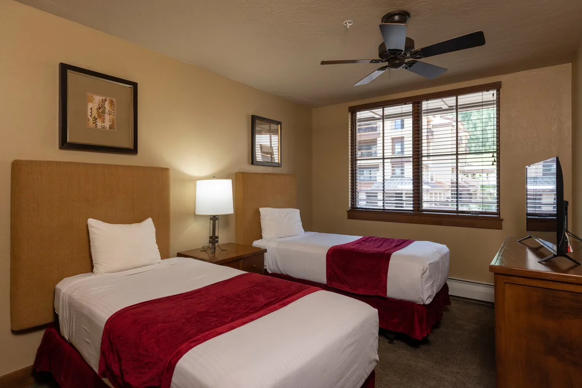 A comfortable twin bedroom with two neatly made twin beds, a nightstand topped with a lamp, a window with blinds, and a television. The setting is cozy and perfect for kids, friends, or families seeking a restful night's sleep.