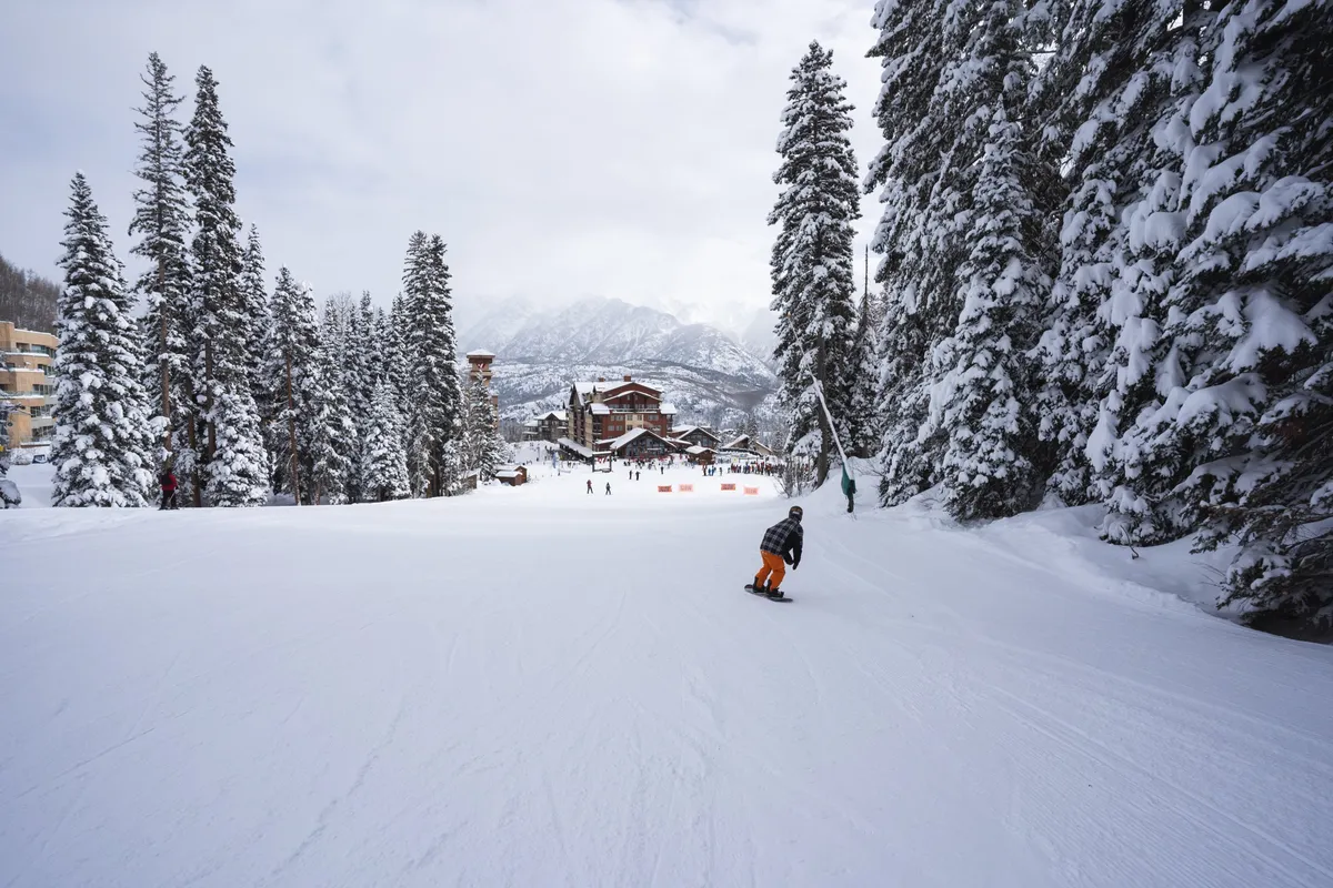 This lively exterior scene captures a snow-covered mountain landscape bustling with skiers and snowboarders near the mountain lodge, surrounded by pine trees and brilliant winter light. The image conveys the energetic atmosphere of outdoor winter activities and the close proximity of the lodge to the slopes.