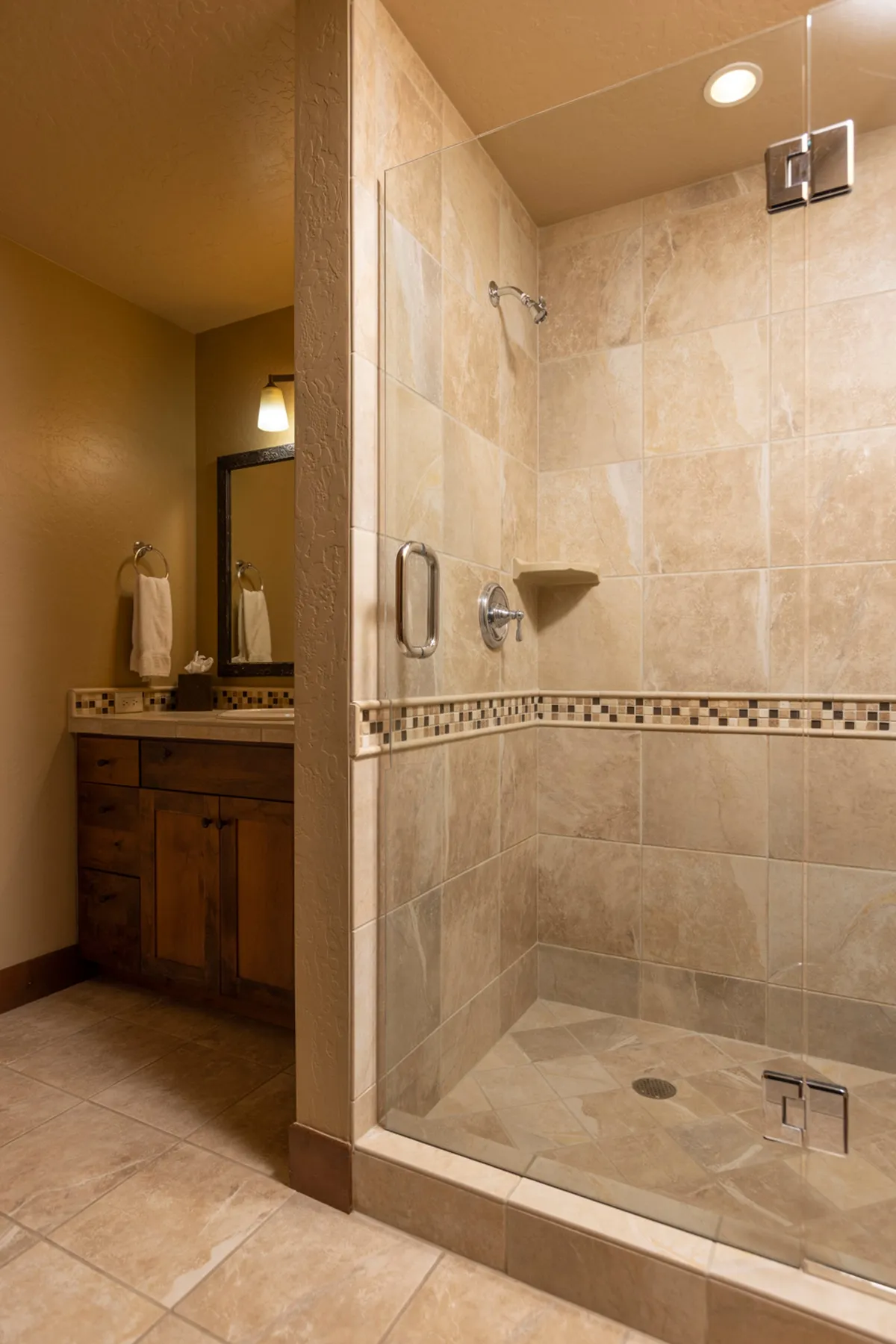 A bright, modern bathroom with a glass shower enclosure, wooden vanity, and a decorative tile border. The scene evokes a tranquil moment of solitude and refreshment, suggesting a private, rejuvenating experience.