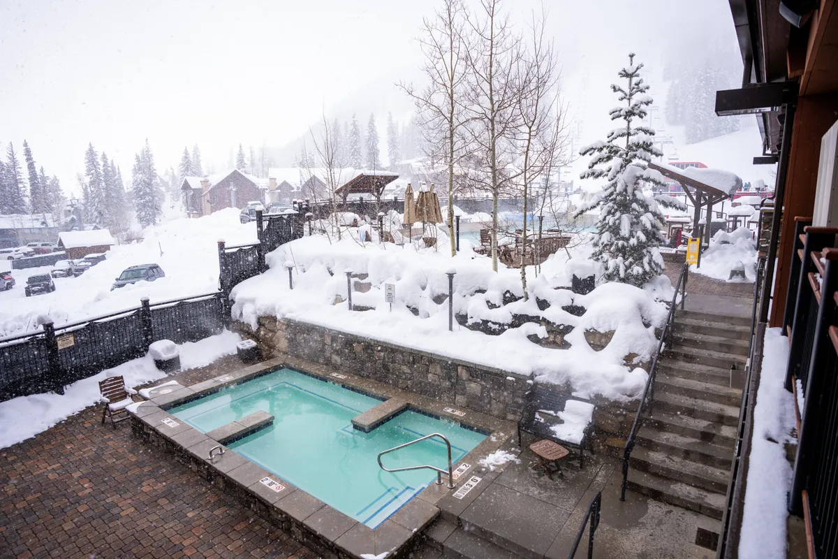 A tranquil outdoor hot tub scene set against a snowy landscape and pine trees, with families or friends relaxing and sharing laughter. The lodge staircase is visible, emphasizing easy access and communal enjoyment in a scenic mountain setting.