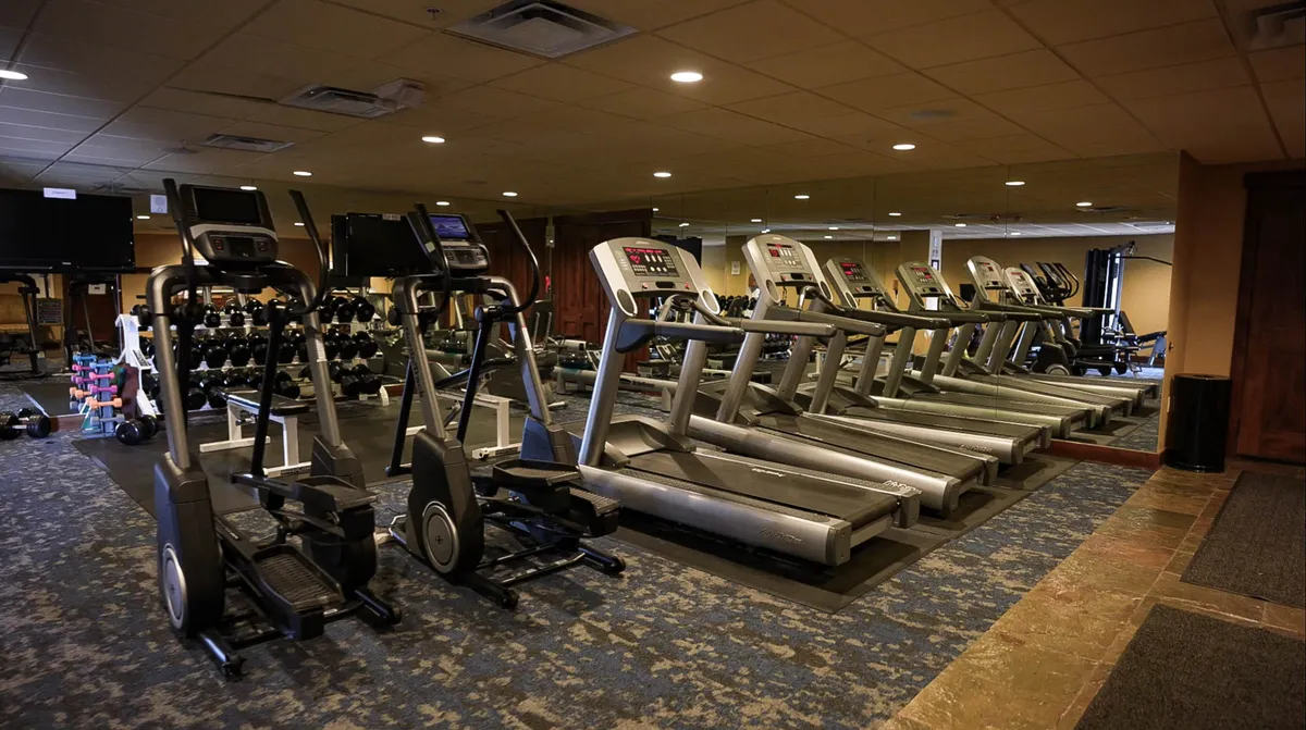 A modern fitness center interior equipped with treadmills, ellipticals, a dumbbell rack, and mirrors. The space exudes an energetic, motivational atmosphere ideal for personal workouts, group classes, or achieving fitness goals while on vacation.