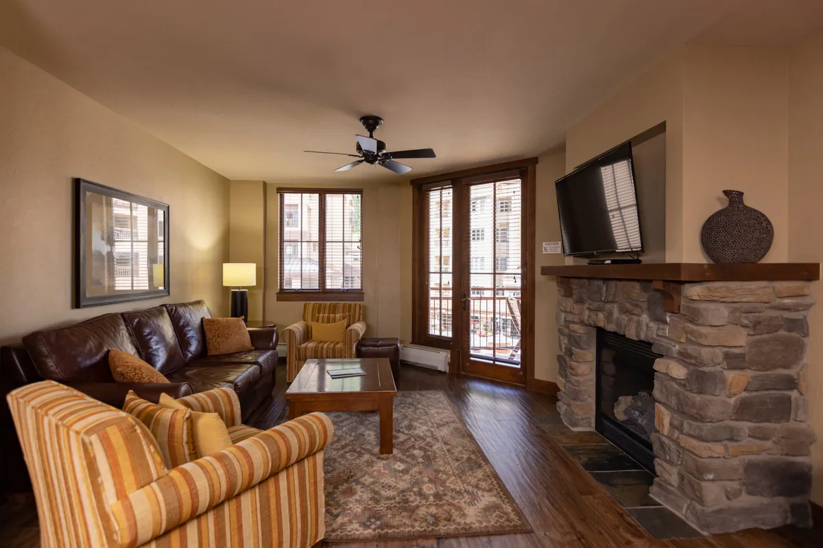 A cozy living room centered around a stone gas fireplace, with plush sofas and a coffee table arranged for conversation. Large windows let in ample natural light and offer picturesque alpine views. The inviting setup is ideal for unwinding after a day outdoors, and the warmth of the fire adds to the gathering-friendly atmosphere.