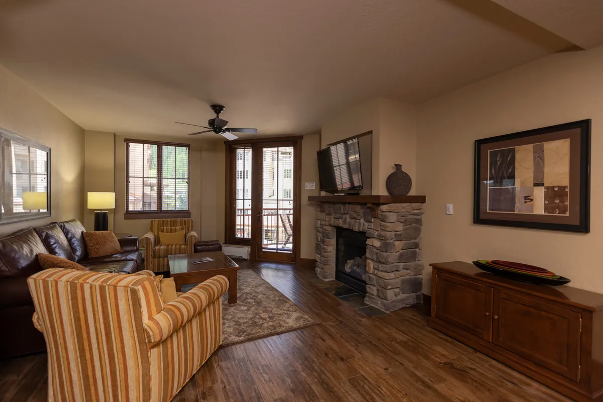 The image showcases the condo's inviting living area, featuring a cozy fireplace, plush sofa seating arranged for conversation, and large windows that flood the space with natural light. The windows offer glimpses of mountain scenery, hinting at stunning views and a balcony perfect for watching sunsets or enjoying morning coffee. The overall mood is warm, comfortable, and sociable, ideal for relaxing after a day on the slopes or gathering with family and friends.