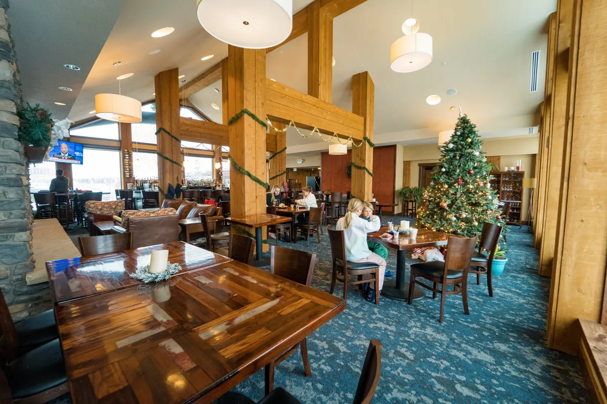 A festive dining area with large windows framing alpine scenery, dining tables set for groups, and a decorated Christmas tree enhancing the atmosphere. The scene hints at lively gatherings, celebrations, and plenty of space for shared meals and socializing.